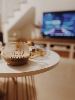 A warm and inviting living room coffee scene with a 'Good Morning' mug and a blurred TV in the background.