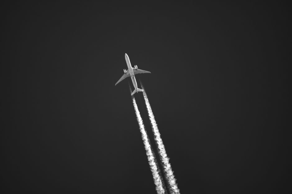 A dramatic black and white image of a jet and contrails soaring through a clear sky, captured from below.