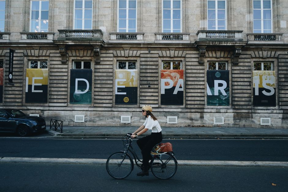 A person bikes past a historic Paris building with artistic window displays.