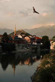 A serene view of an old city by riverside at sunset with mountains in the background and a bird soaring above.
