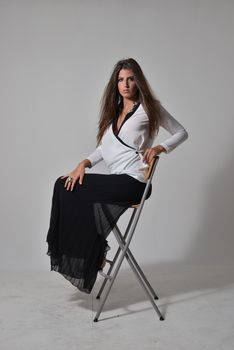 Stylish woman in white blouse and black skirt sitting on a stool in a studio setting.