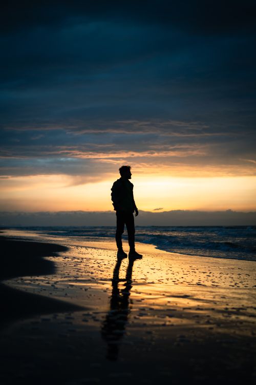 Free A lone man walking along the shoreline of an ocean during sunset, creating a striking silhouette against the vivid sky. Stock Photo