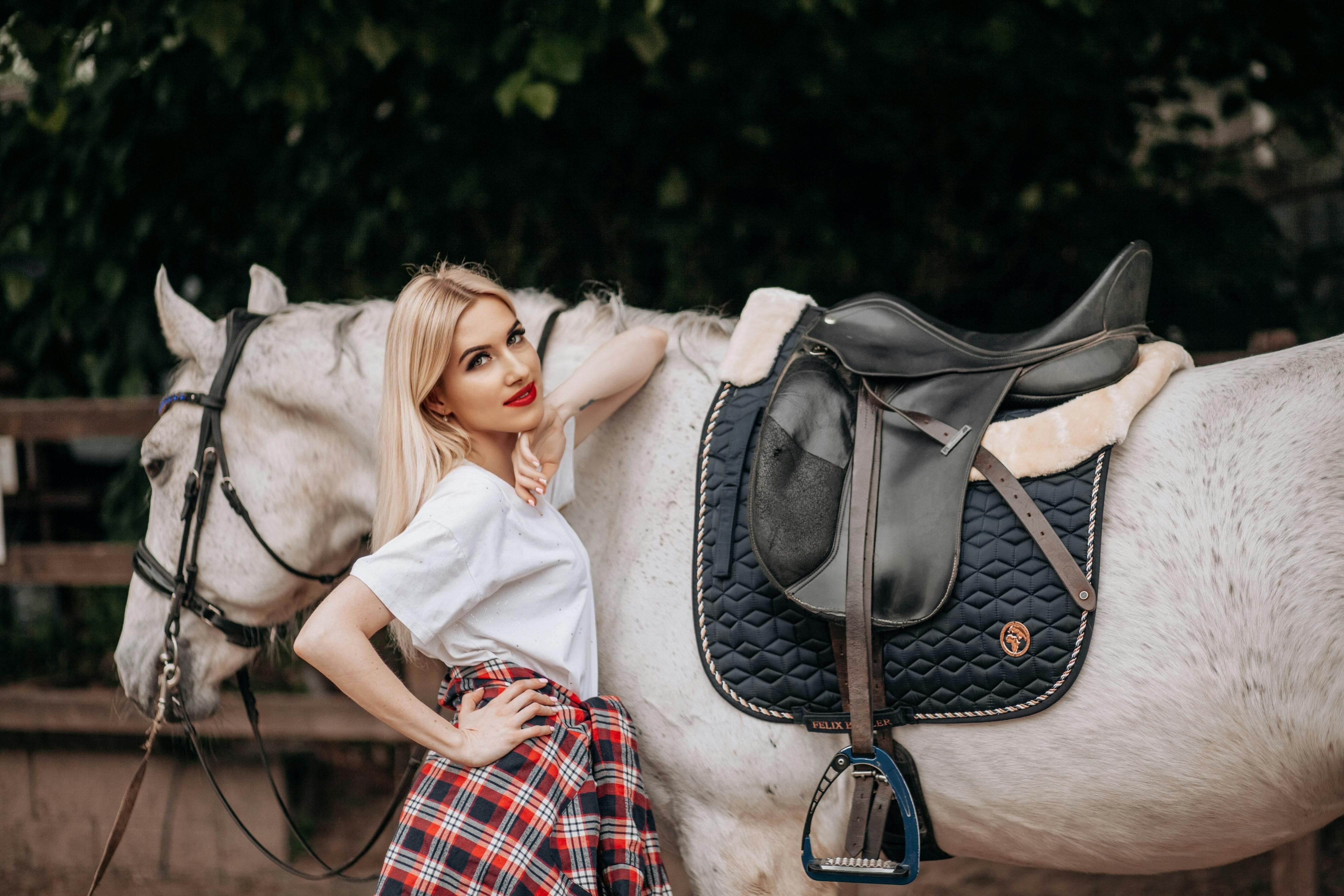 A woman stands beside a white horse with saddle, smiling in an outdoor setting.