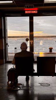 An elderly man sits in Bostancı ferry terminal with a view of the sea, reflecting solitude.