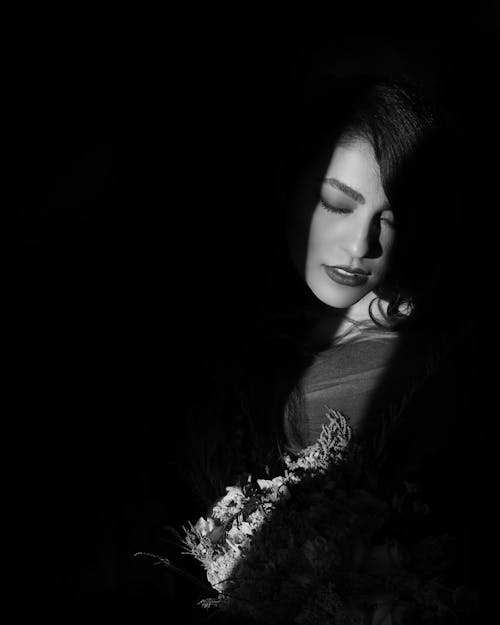 Light and Shadow Photo of Woman Inside Dark Room Posing With Her Eyes Closed While Holding a Bouquet of Flowers