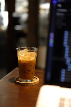 Chilled iced coffee in a glass beside a blurred laptop screen in a cozy indoor cafe.