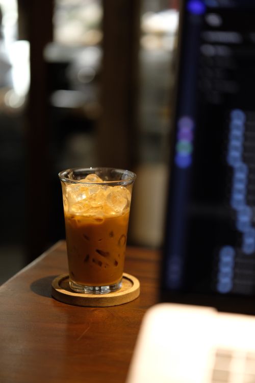 Free Chilled iced coffee in a glass beside a blurred laptop screen in a cozy indoor cafe. Stock Photo