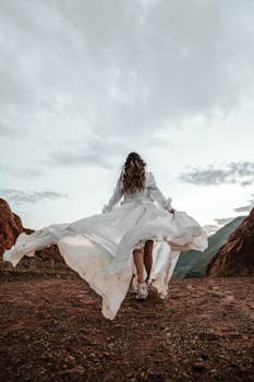 在多云的天空下，一名身穿飘逸白裙的女子走在岩石山间小路上，留下一个背影