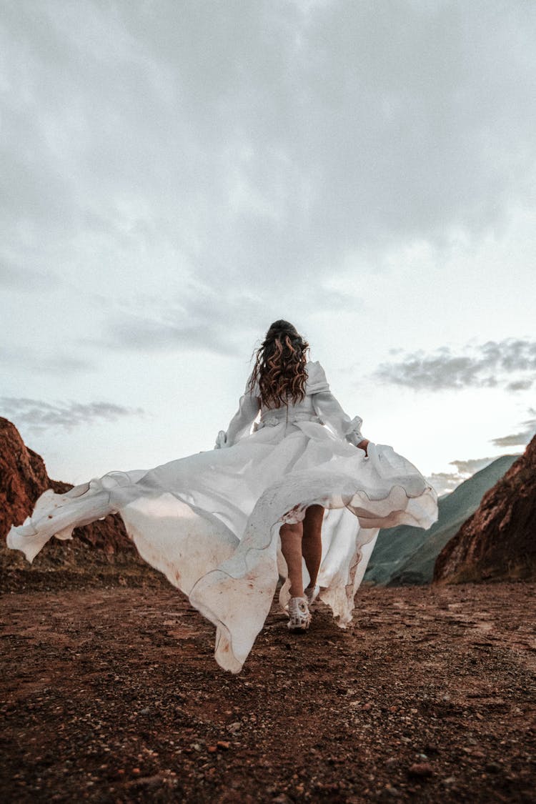 Back View Photo Of Woman In White Dress Walking On Muddy Path