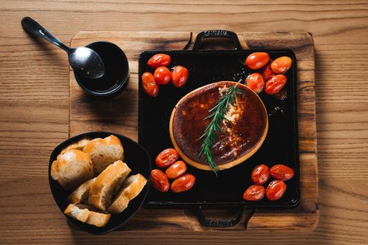 Flat lay of baked cheese with cherry tomatoes, fresh bread, and a rosemary garnish on a cutting board.