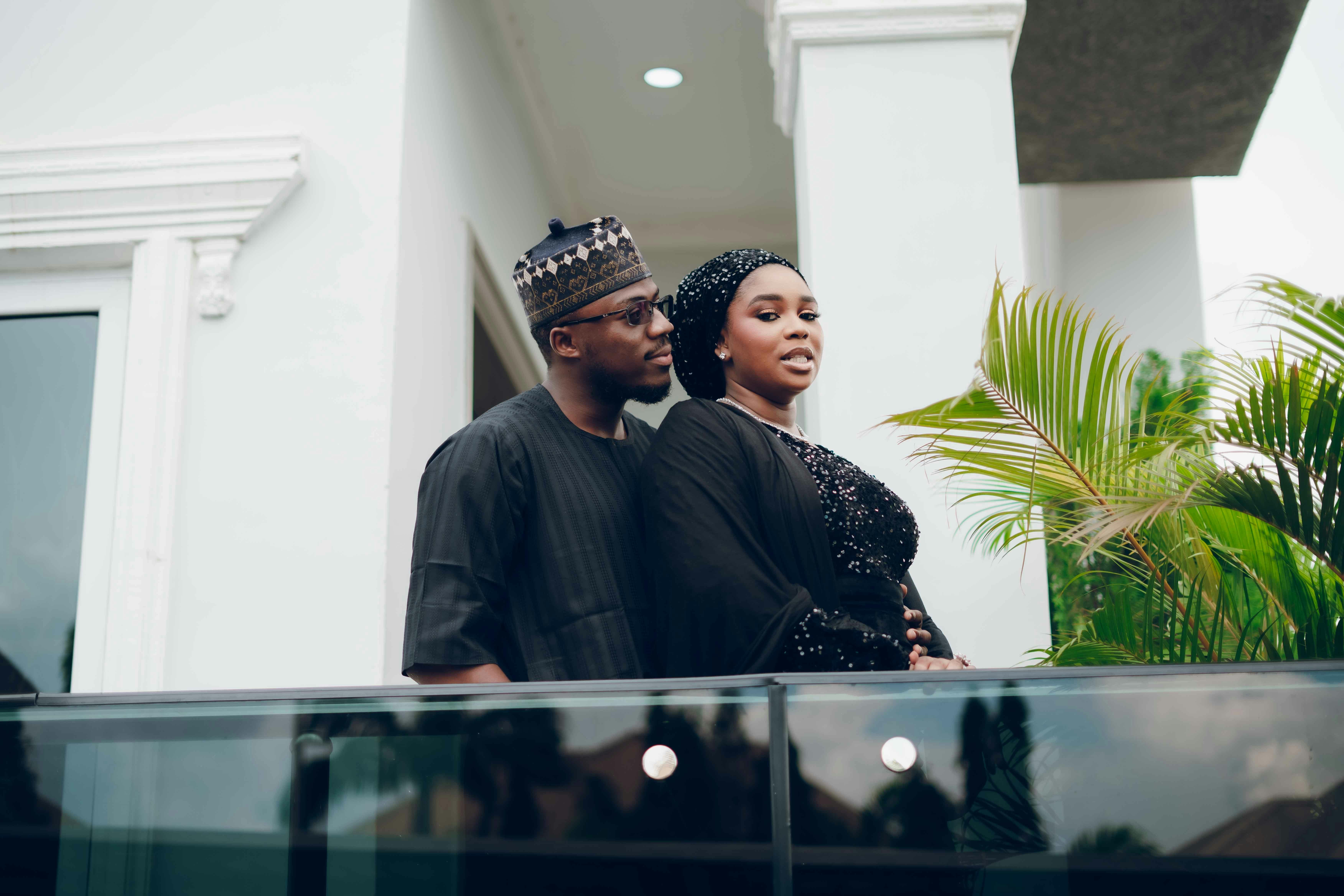 A couple stands elegantly on a balcony, dressed in traditional African attire, with lush green plants in the background.