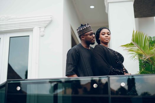 A couple in black traditional attire poses elegantly on a modern balcony.