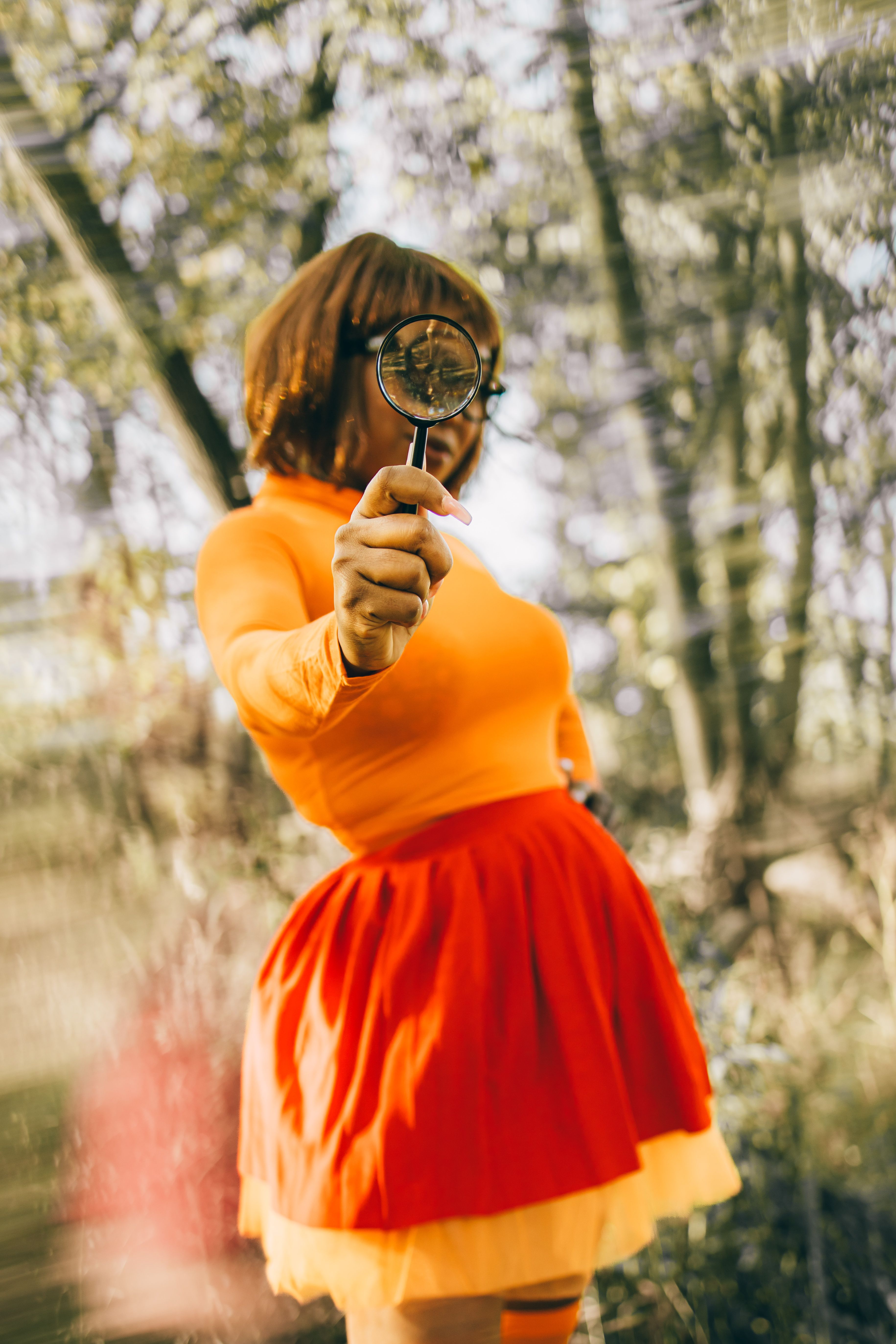 A person in cosplay holding a magnifying glass, set against a vibrant outdoor backdrop.
