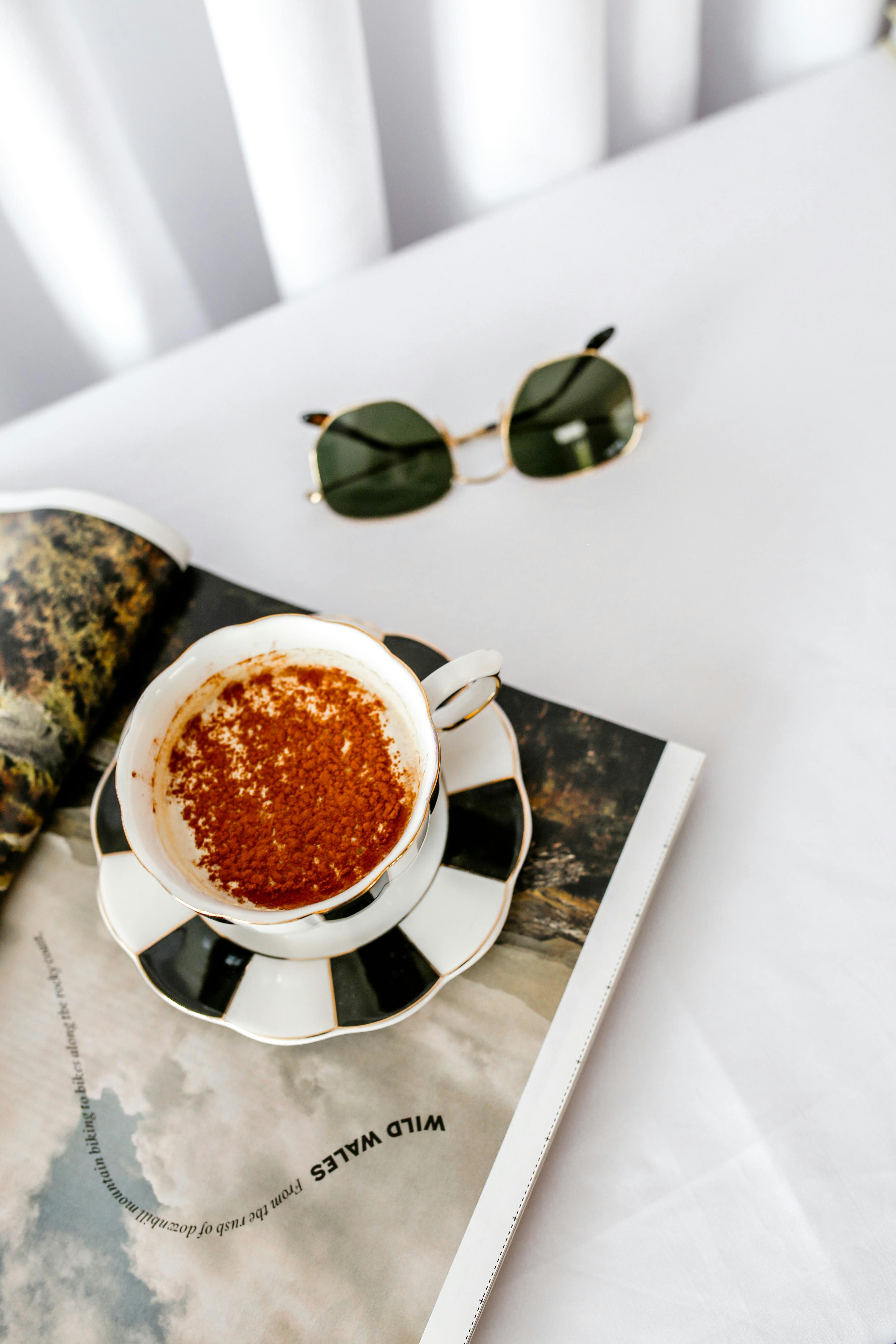 Aesthetic coffee scene featuring a cappuccino, open magazine, and sunglasses on a white tablecloth.