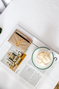 A stylish flat lay of a cappuccino, magazine, and luxury wallet on a white table.