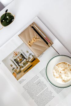 A flat lay image with a magazine, sunglasses, cappuccino, and designer cardholder on a white surface.