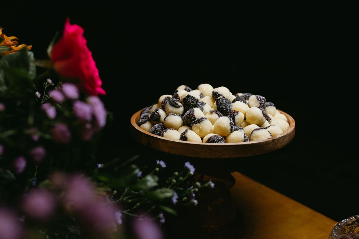 A wooden bowl filled with sugar-coated confections surrounded by vibrant flowers in a dim setting.