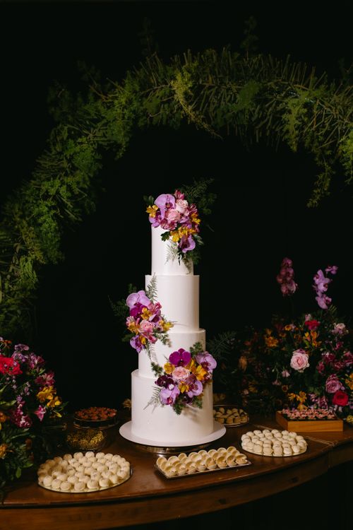 Free A three-tiered wedding cake adorned with vibrant flowers, surrounded by desserts against a lush green arch at night. Stock Photo