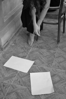 Black and white image of a woman sitting with papers on the floor in an artistic setting.
