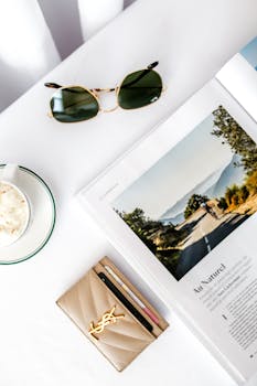 Minimalist flat lay showcasing sunglasses, magazine, coffee, and a wallet on a white surface.