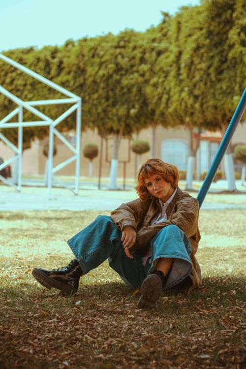 Free Young woman sitting casually on grass, leaning against a pole in a park setting. Stock Photo