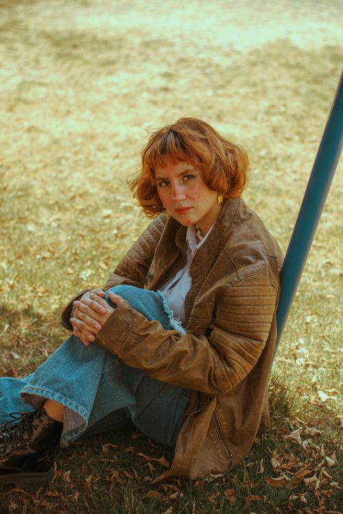 Free A young woman with red hair sits on the grass in a brown jacket, appearing contemplative. Stock Photo