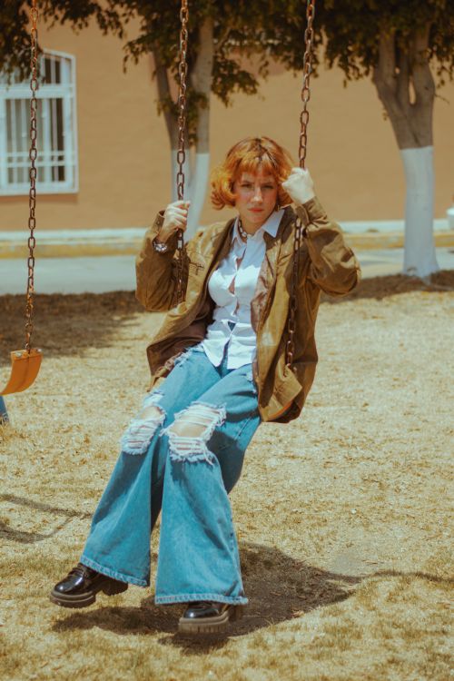 Free Stylish young woman with red hair enjoys a swing in a sunny outdoor setting, wearing ripped jeans and a jacket. Stock Photo