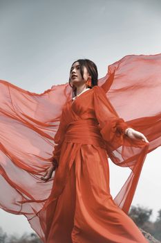 A woman in a flowing red dress poses gracefully outdoors with a dramatic sky backdrop.