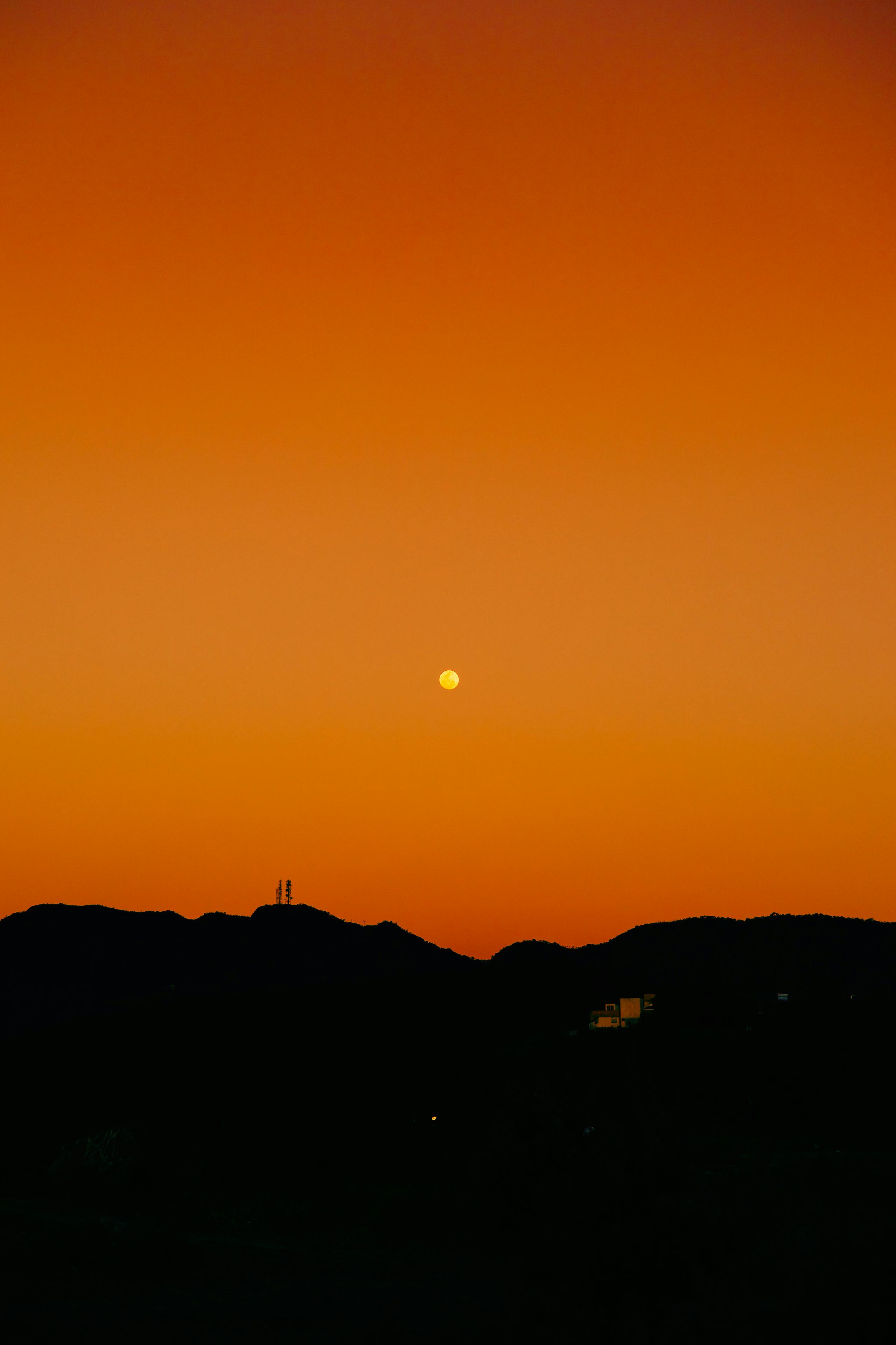 Captivating orange sunset with full moon over silhouetted mountains.