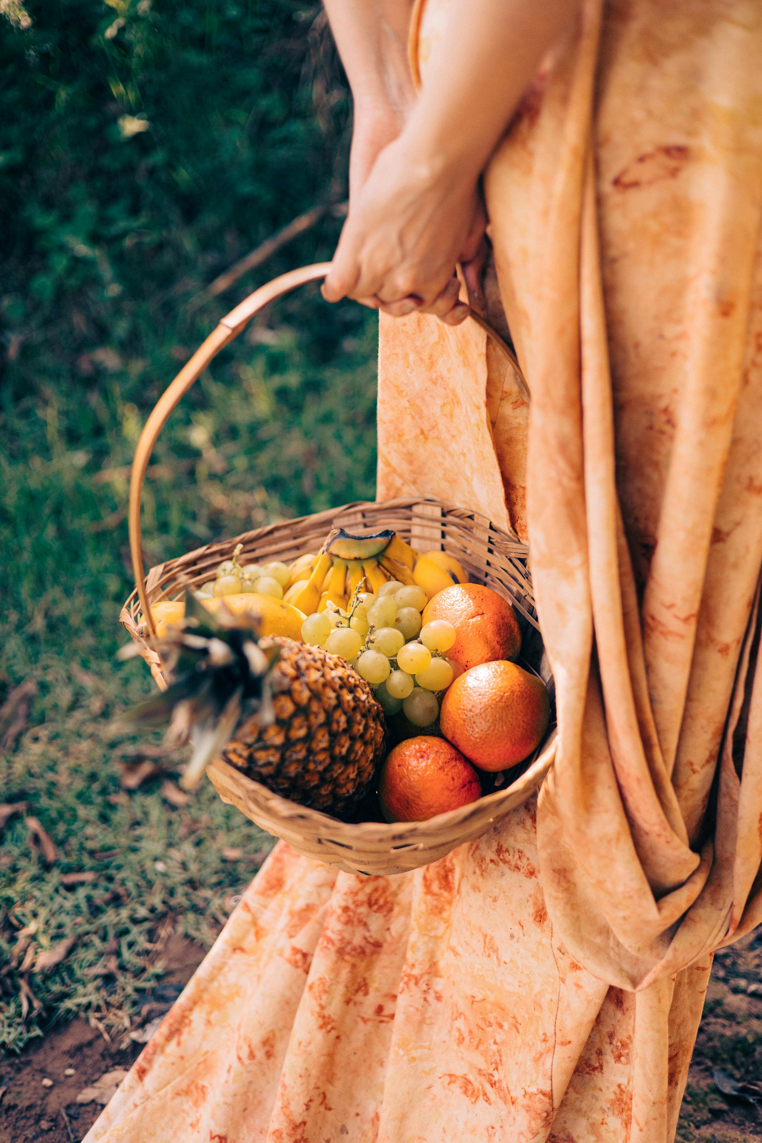 A woman in an orange dress holds a wicker basket filled with tropical fruits like pineapple and oranges outdoors.