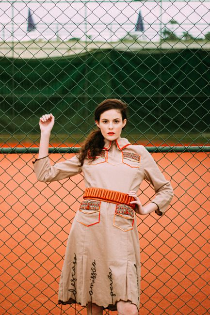 Stylish woman in a retro dress posing confidently against a tennis court fence.