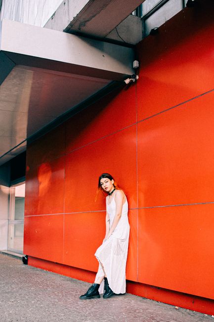 Stylish woman in a white dress and black boots poses by a vibrant red wall in an urban setting.