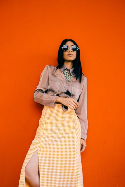 Stylish woman wearing sunglasses and fashionable outfit poses against a vibrant orange wall.