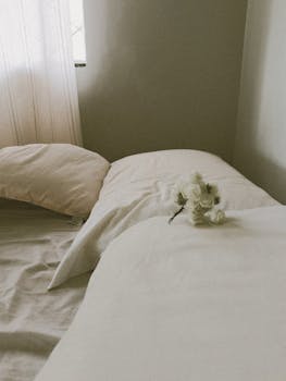 A serene bedroom scene with soft pillows and white flowers on a bed, bathed in gentle natural light.