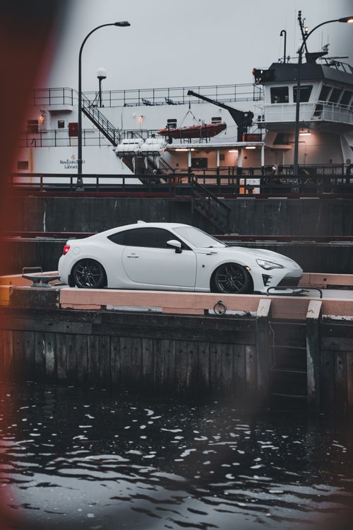 Free White sports car parked by shipyard, moody clouds overhead, water reflection. Stock Photo