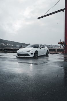 A modern white sports car parked on a wet, reflective pavement in an industrial area with cloudy skies.