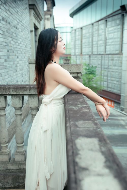 Free A serene woman in a white dress gazing from a balcony in a historic urban setting. Stock Photo