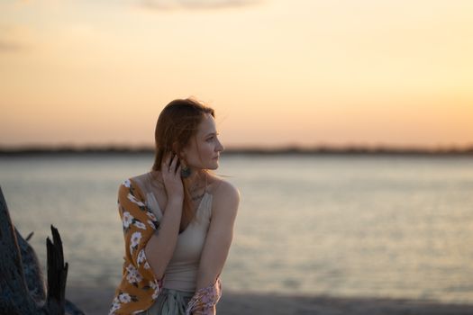 A woman with floral shawl looks thoughtfully out to a serene beach sunset.