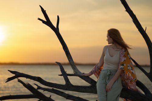 Free A woman standing on driftwood by the water, gazing at the sunset. Stock Photo