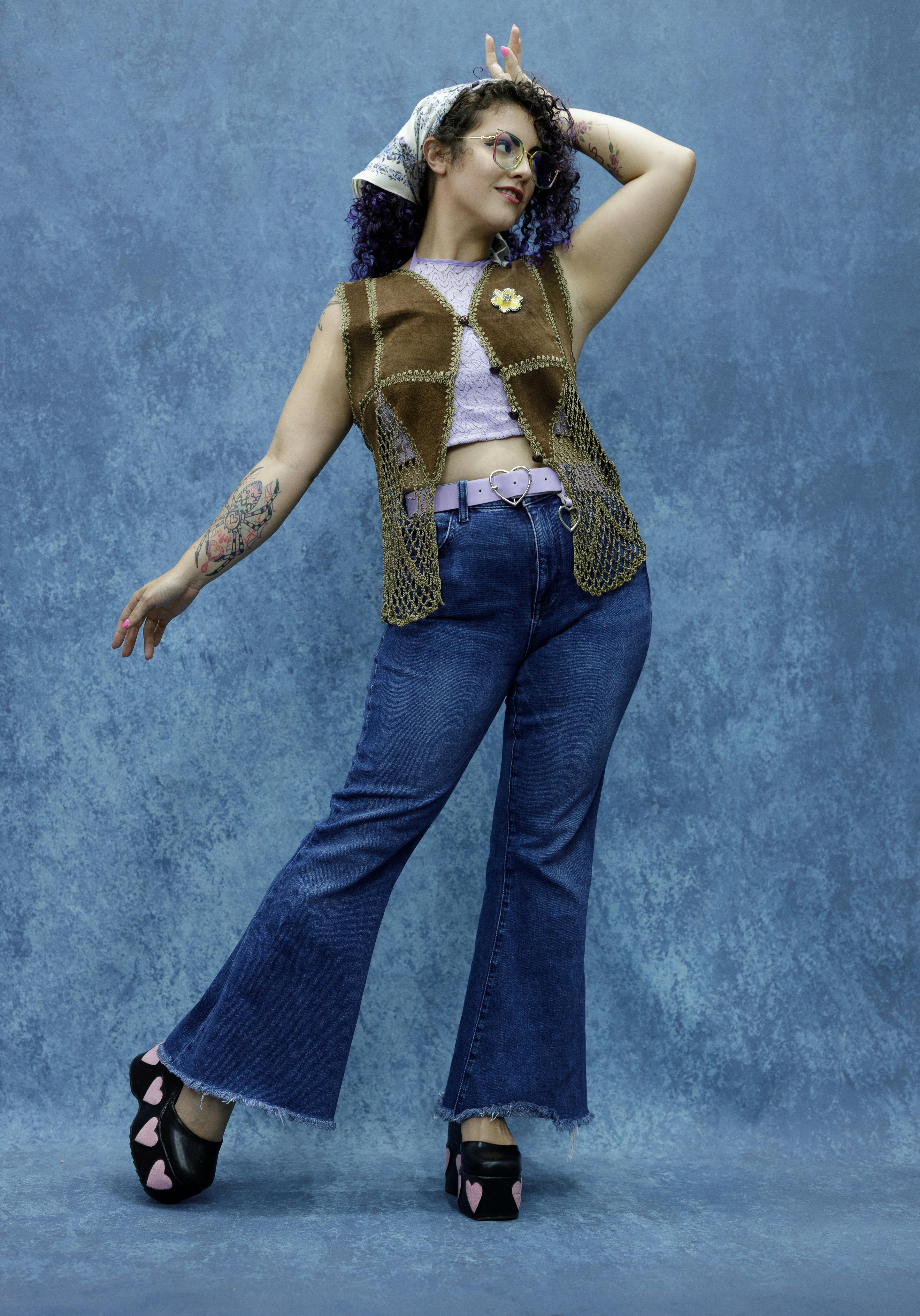 Trendy woman strikes a playful pose in retro 70s fashion with high-waisted jeans and crochet vest.
