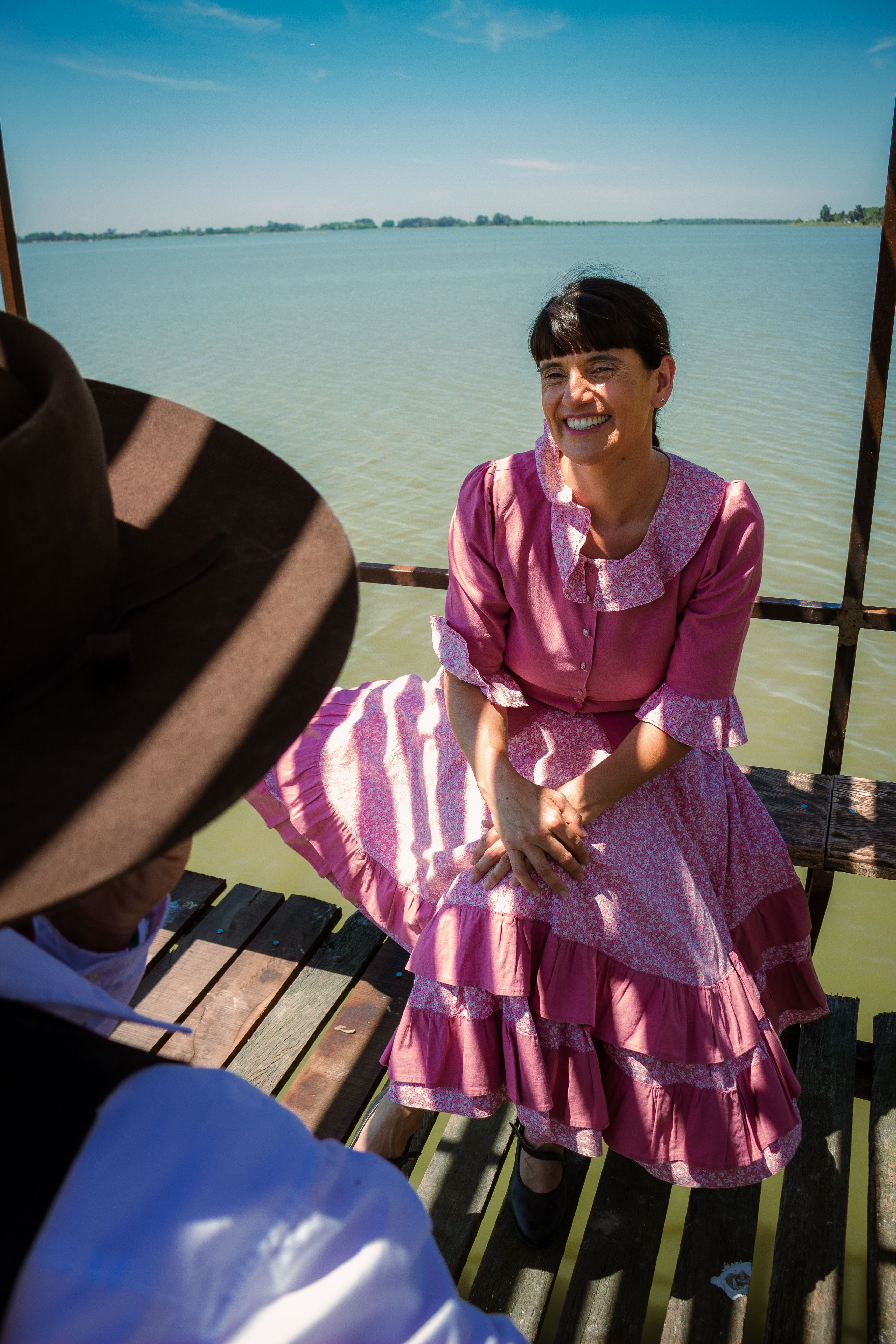 A smiling woman in a pink traditional dress enjoying a sunny day by the lakeside.