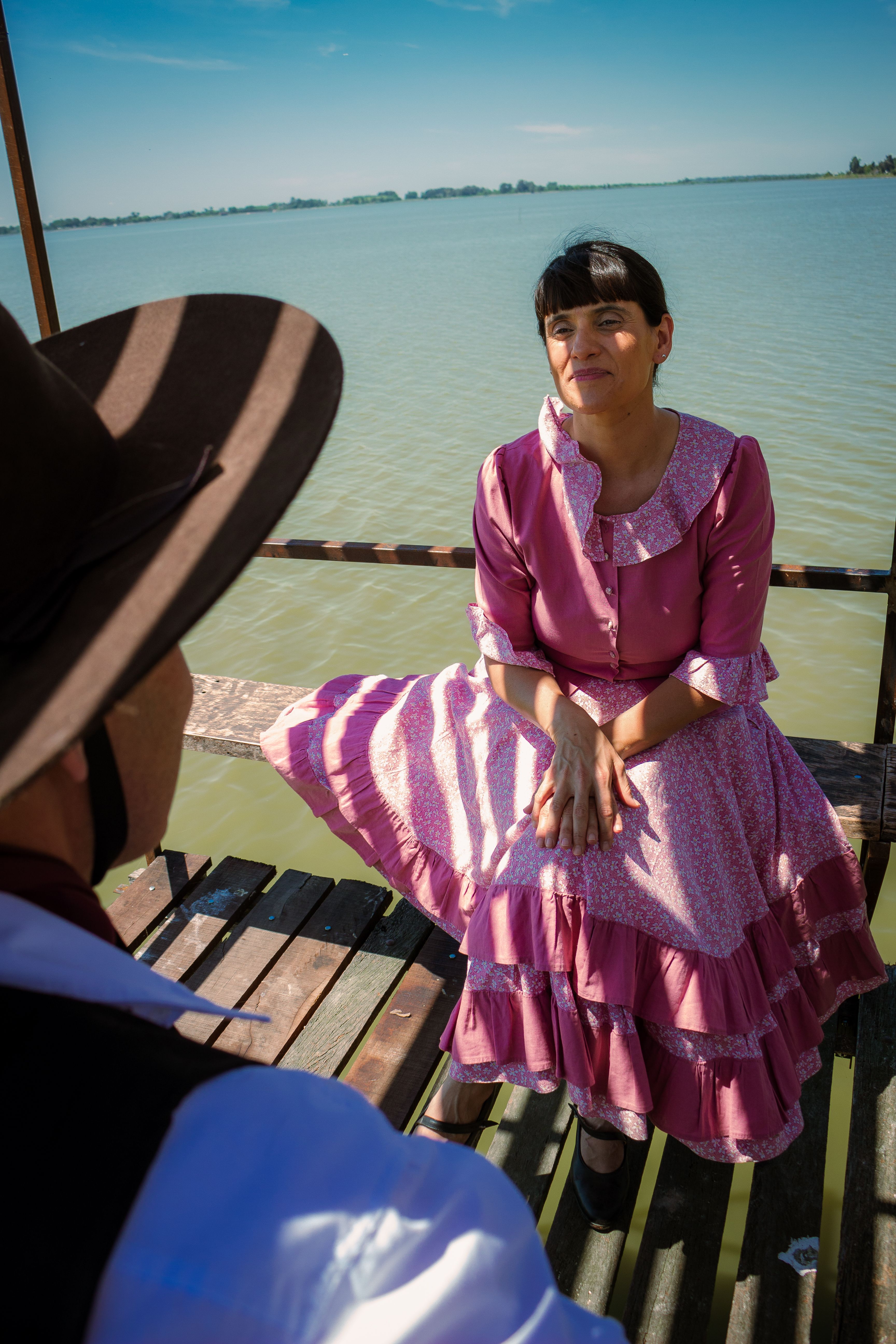 A man and woman in vintage clothing enjoying the outdoors by a lake pier on a sunny day.