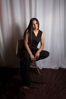 Portrait of a stylish woman sitting on a chair against a draped white curtain.
