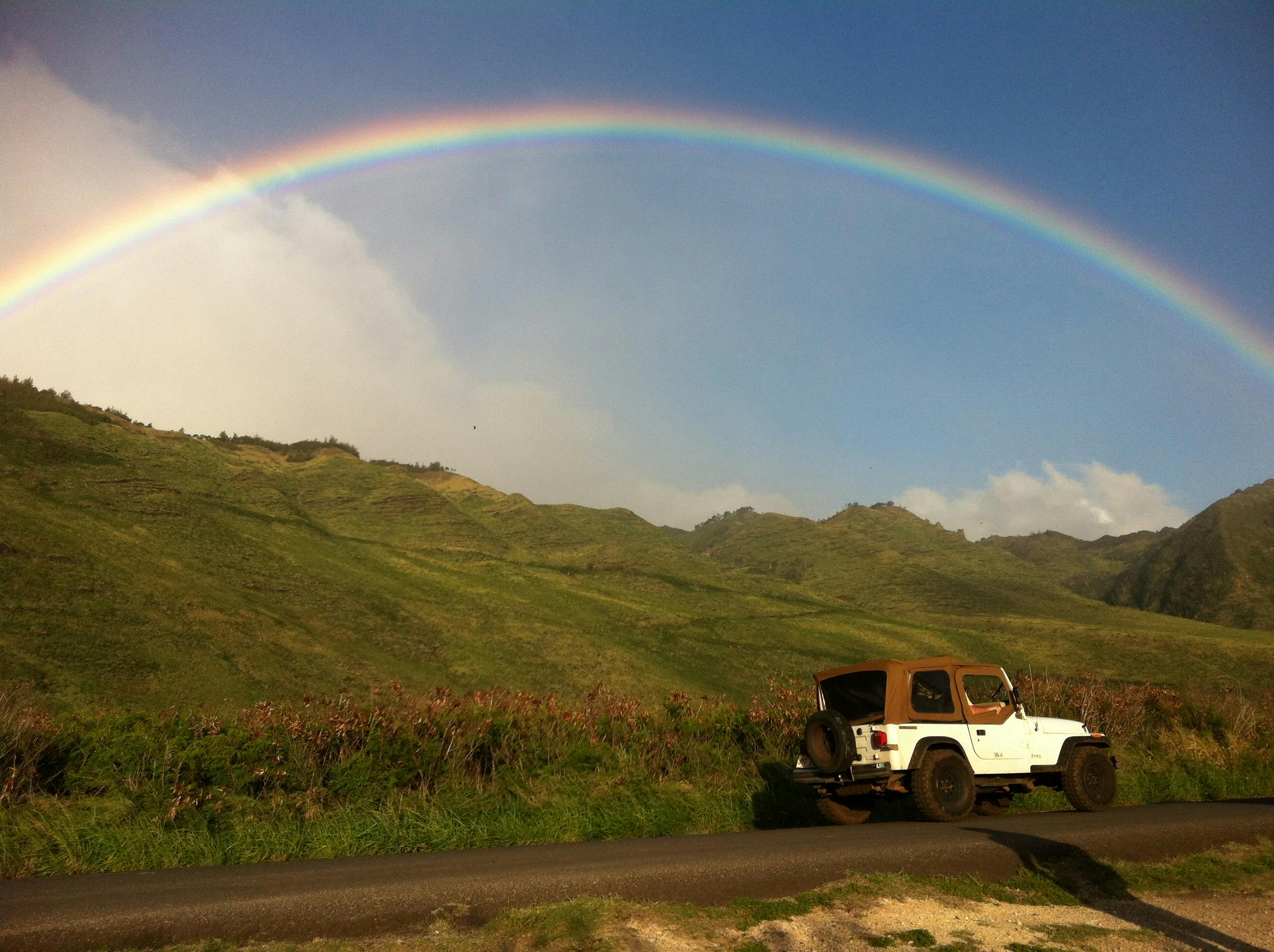 Stunning view of a rainbow over lush hills with a Jeep on a road in Hawaii.