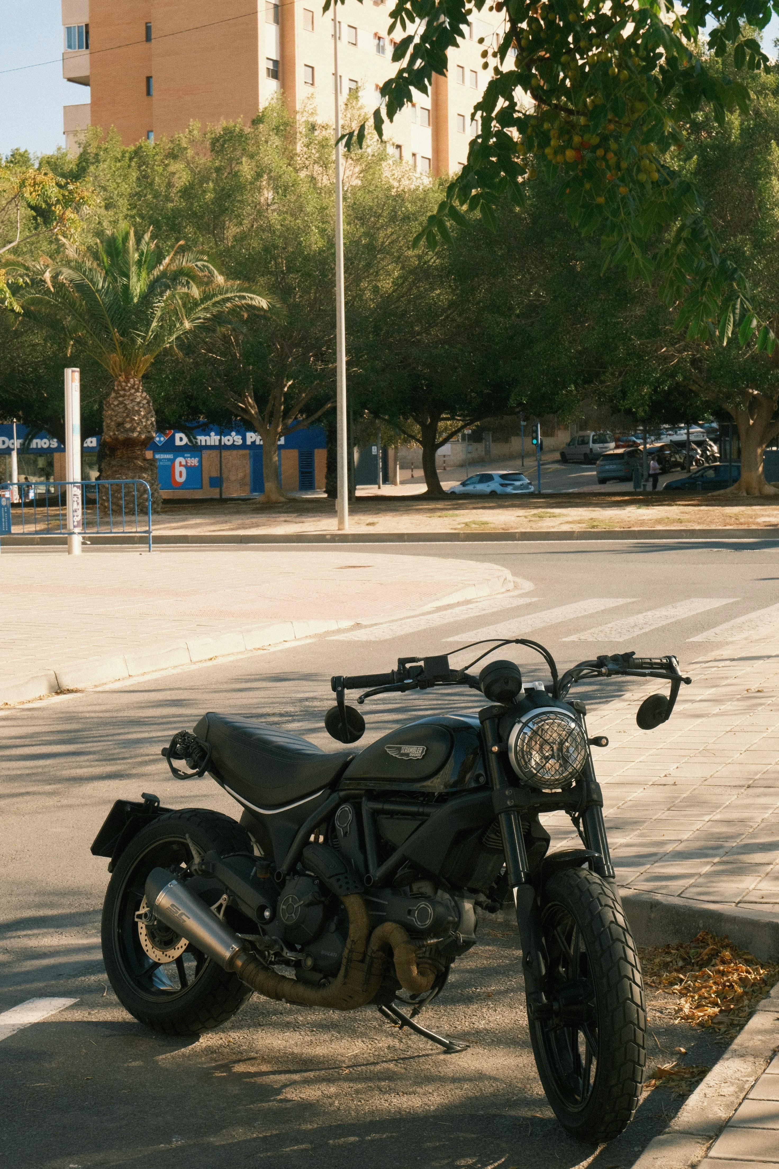 Free A classic motorcycle parked in a sunny urban setting with buildings and trees. Stock Photo