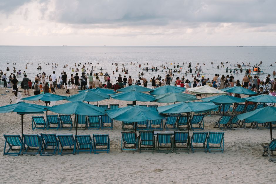 A lively beach scene with blue umbrellas, chairs, and people enjoying the sea.