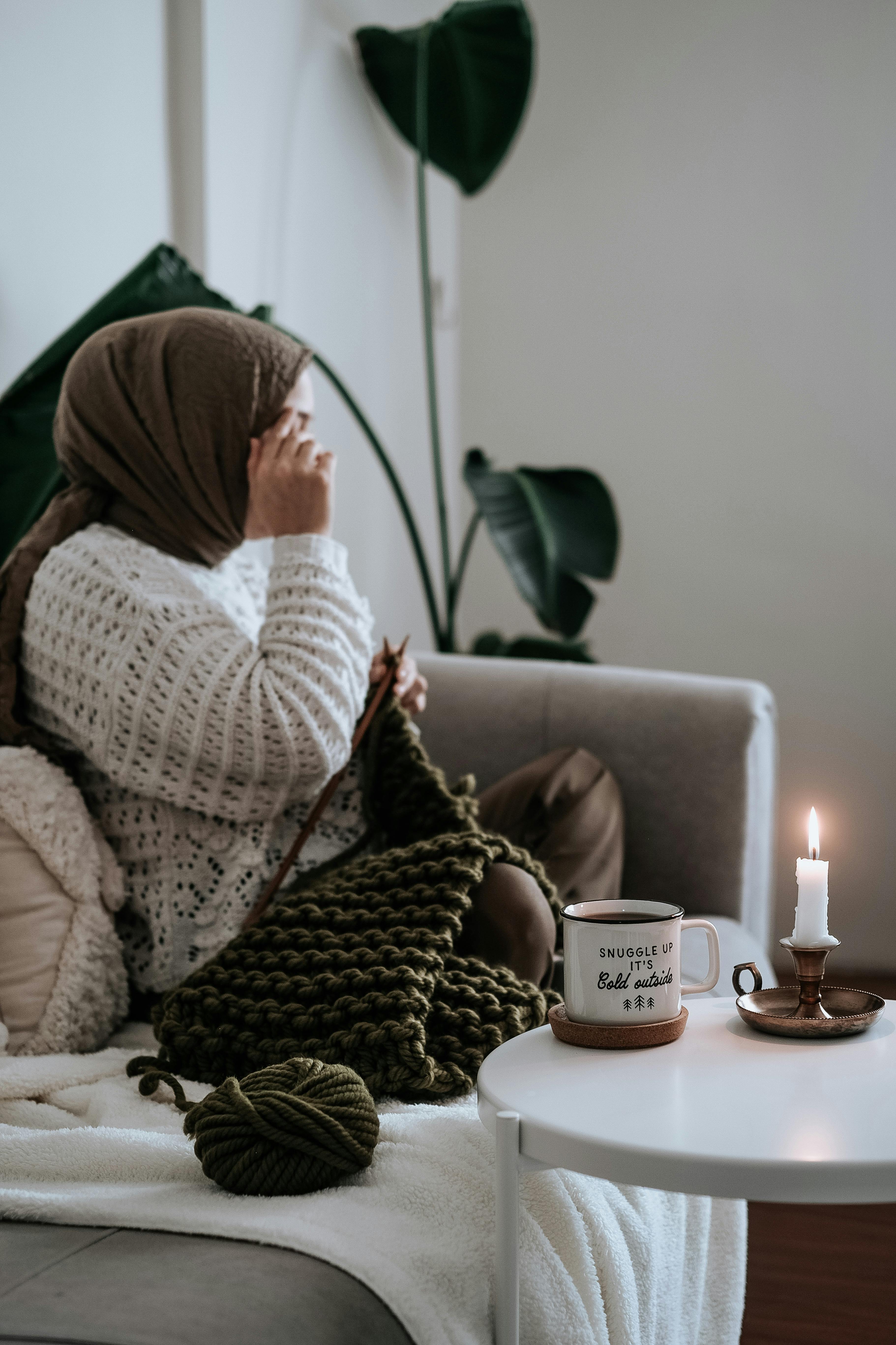Woman knitting on couch with candle, coffee, and plant. Cozy indoor setting.
