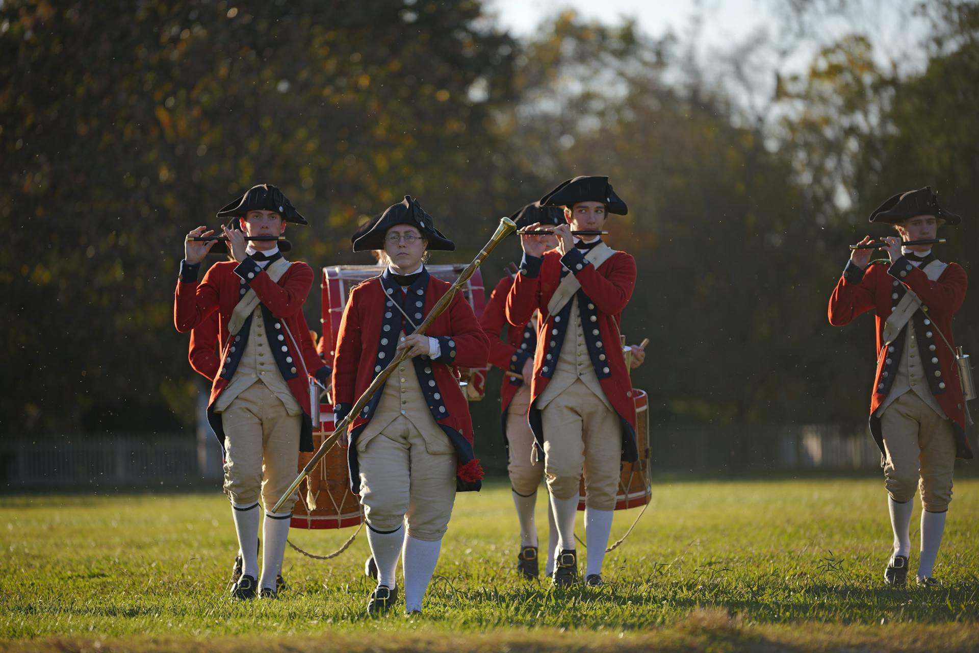 Colonial soldiers in red uniforms perform in a historical reenactment outdoors.