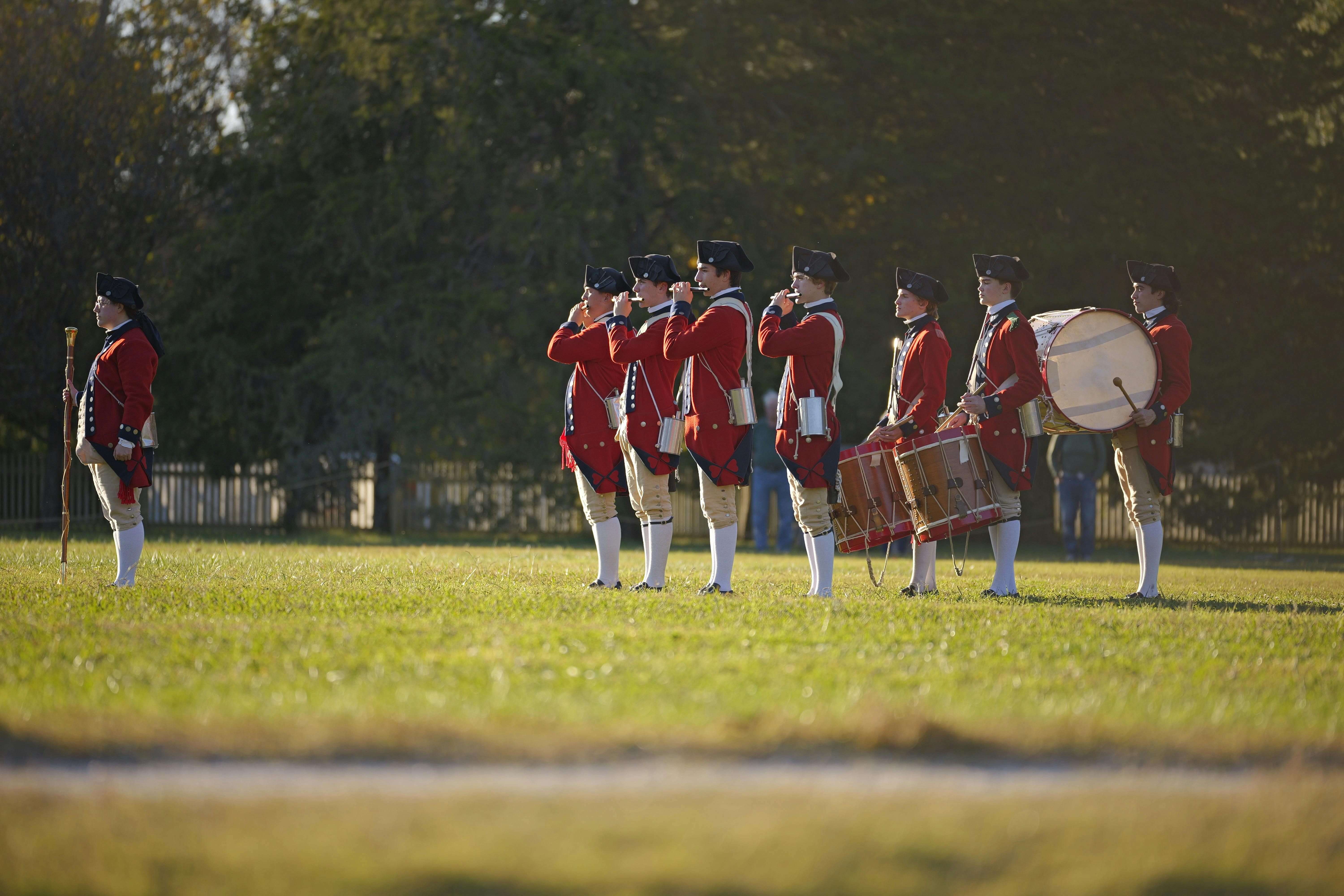 Colonial-era military band reenacting history in open field.