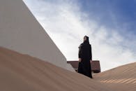 这是一张低角度镜头拍摄的照片，一名身穿传统长袍的女子站在晴朗天空下的沙漠沙丘上